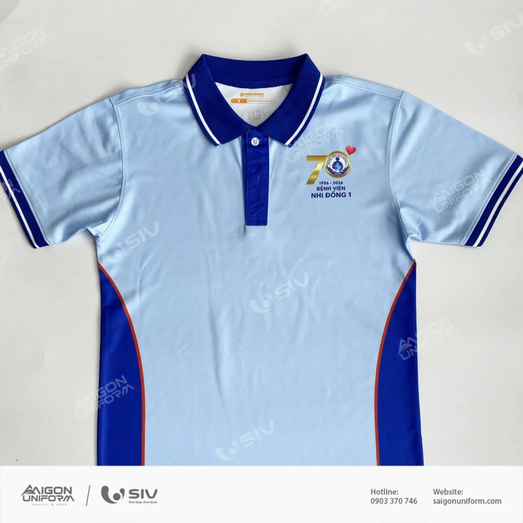 Áo thun đồng phục Bệnh viện Nhi Đồng 1 kỷ niệm 70 năm do Saigon Uniform sản xuất. Vải cá sấu cà phê, bo cổ xanh đậm hai sọc trắng, hông ghép xanh viền cam. Áo polo đồng phục Bệnh viện Nhi Đồng 1 màu xanh dương nhạt, logo kỷ niệm 70 năm 1956 2026 in chuyển nhiệt ở ngực trái